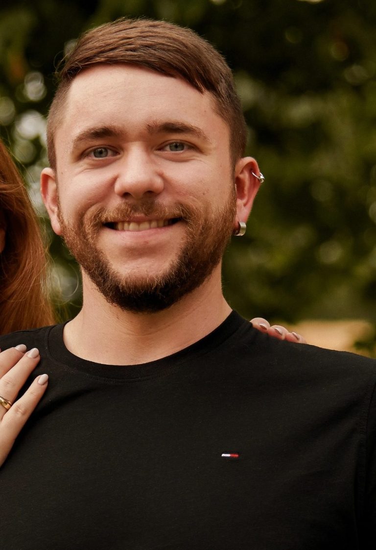 Ein lächelnder Mann mit kurzen Haaren und Ohrringen, trägt ein schwarzes T-Shirt. (Fladiplushies Inhaber)