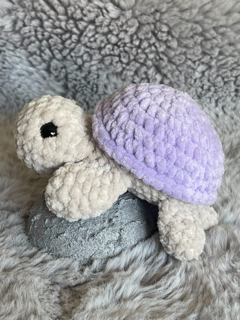 Gehäkelte Schildkröte mit beigem Körper und lila Panzer auf einem grauen Hintergrund.